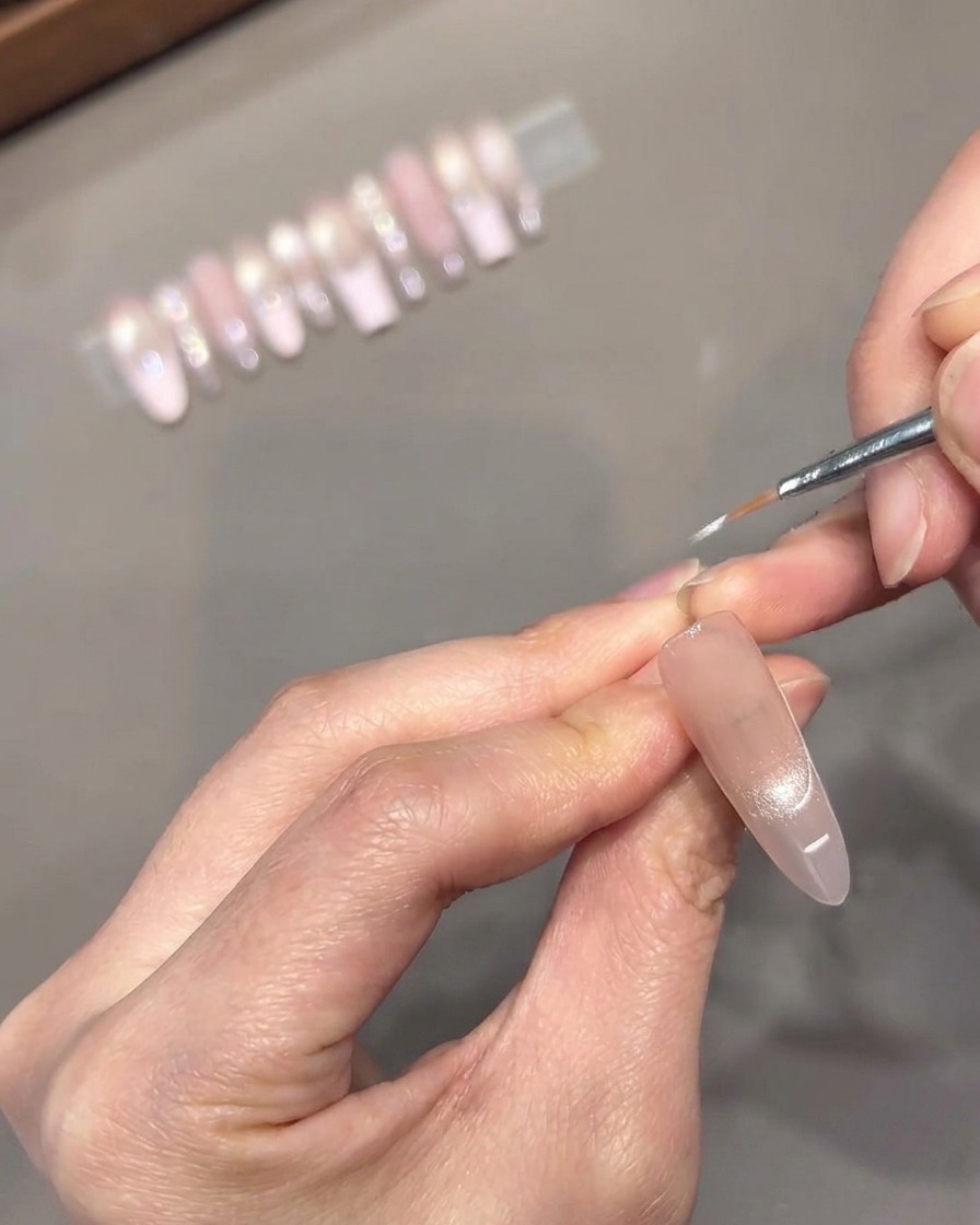 Un pinceau liner fin dessinant une ligne de sourire French rose pâle précise sur la partie supérieure dun ongle.