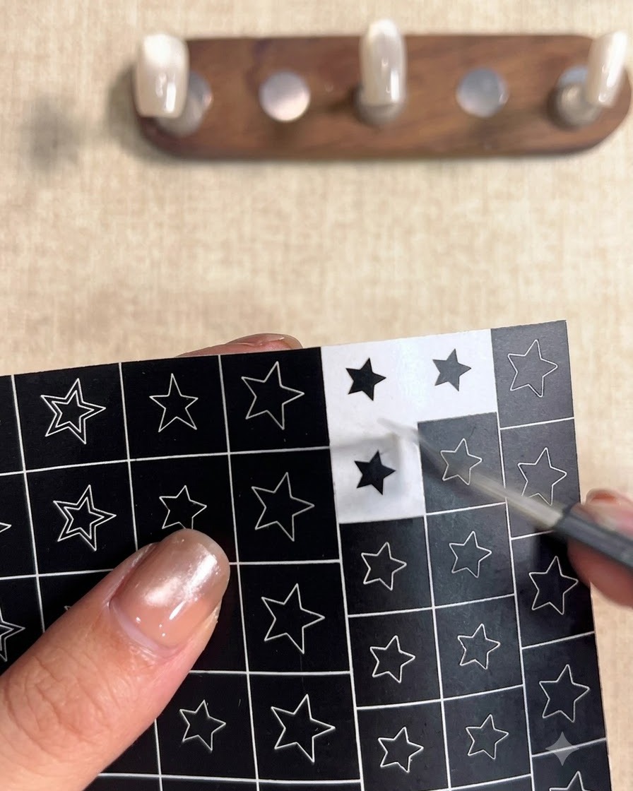 Une main utilisant une pince à épiler pour décoller un autocollant carré noir avec une découpe détoile évidée dune planche de motifs détoiles.