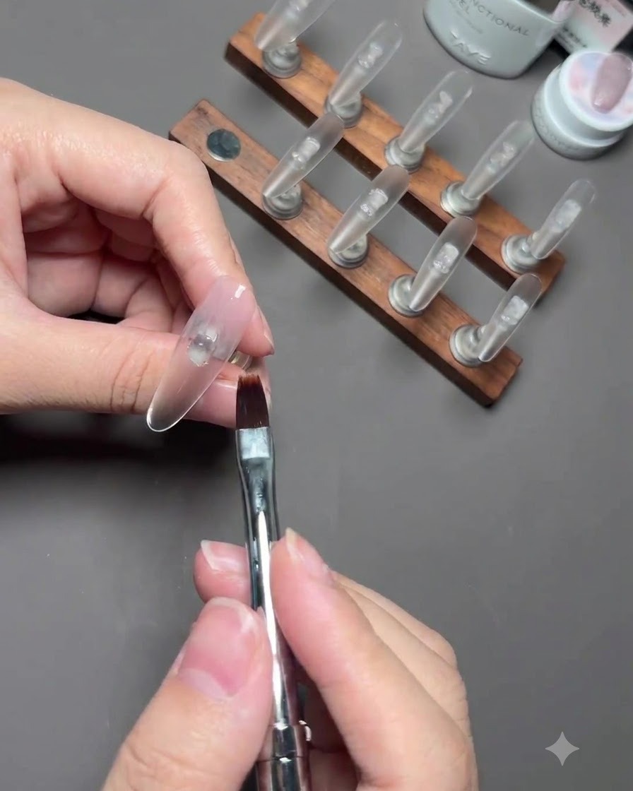 Un pinceau à ongle estompant du gel polish nude diaphane sur longle dun pouce fixé sur un support.