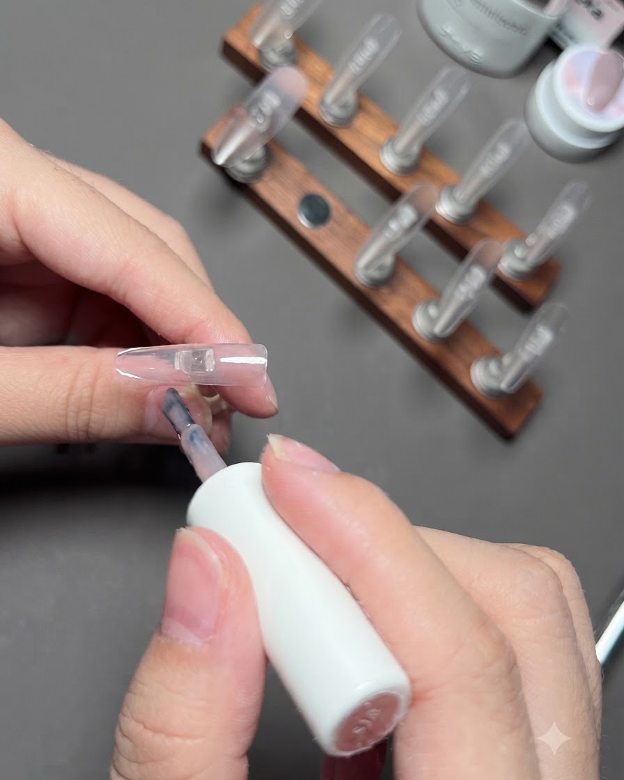 Un pinceau appliquant du gel couleur nude sur longle de lannulaire.