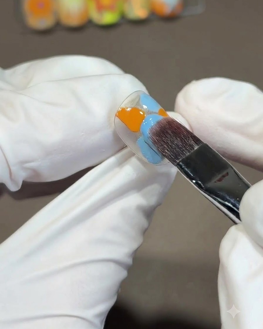 Un grand pinceau carré mélangeant des vernis gel orange et bleu sur une capsule dongle en effectuant un mouvement descendant en forme de S.