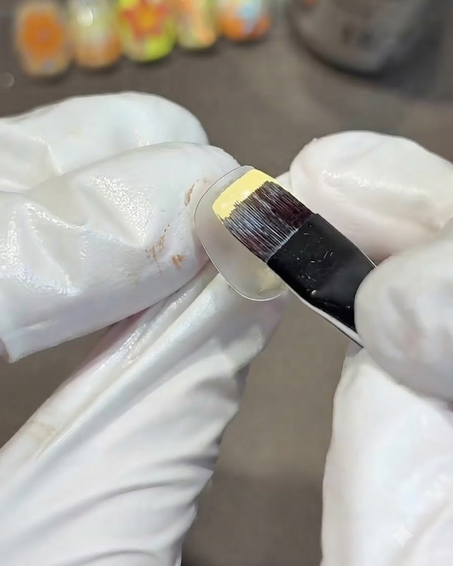 Une main gantée utilisant un pinceau gel plat pour peindre une capsule dentraînement transparente avec un vernis gel jaune uni.
