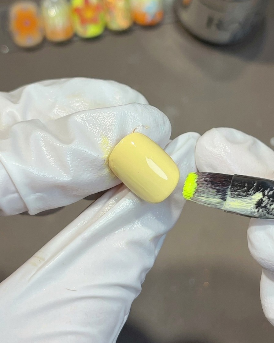 Une main gantée utilisant un pinceau plat pour appliquer du vernis gel vert vif sur le coin dun ongle peint en jaune.