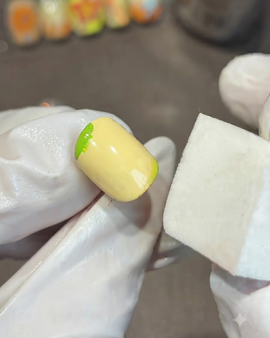 Une main gantée tenant un bloc éponge blanc à côté dun ongle jaune, estompant un dégradé de gel vert sur le coin.