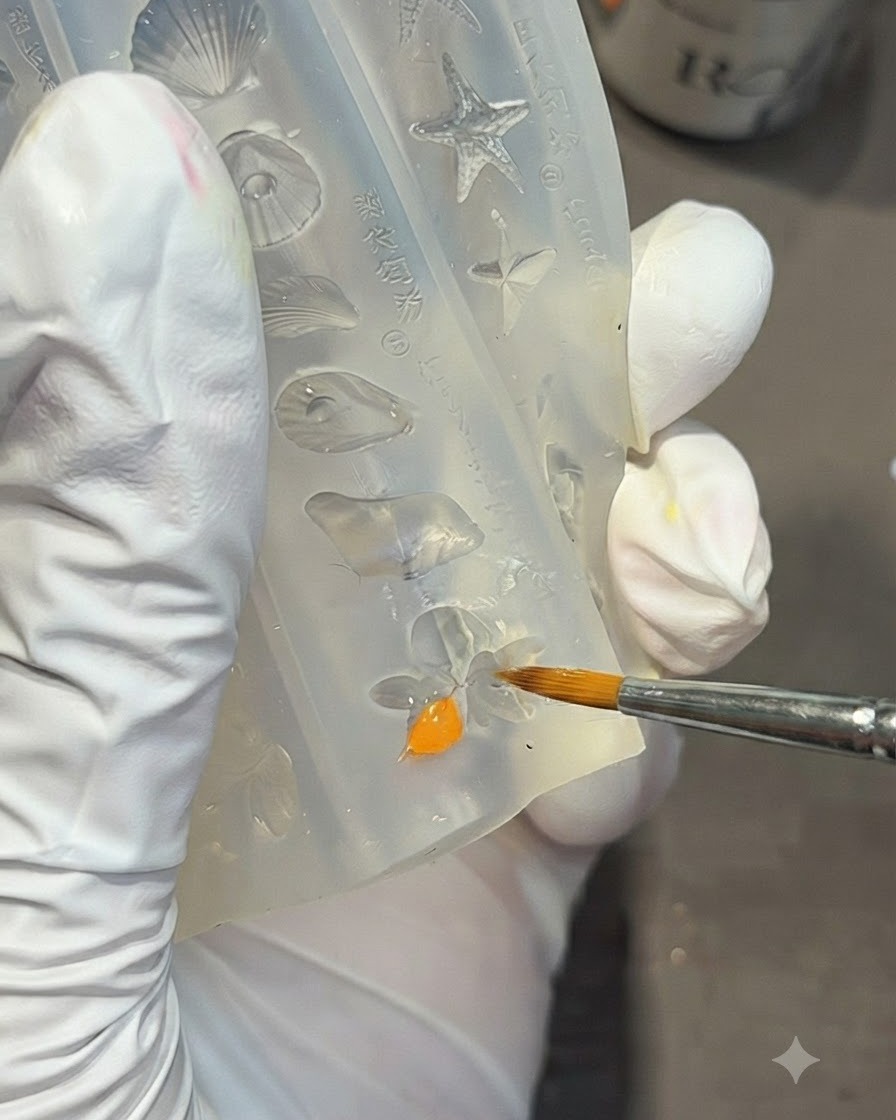Une main gantée utilisant un pinceau de détail pour remplir la forme dune fleur de lys à lintérieur dun moule en silicone transparent avec du gel orange vif.