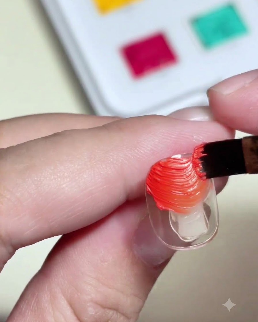 Un pinceau appliquant du gel paint orange et jaune texturé sur une capsule transparente.