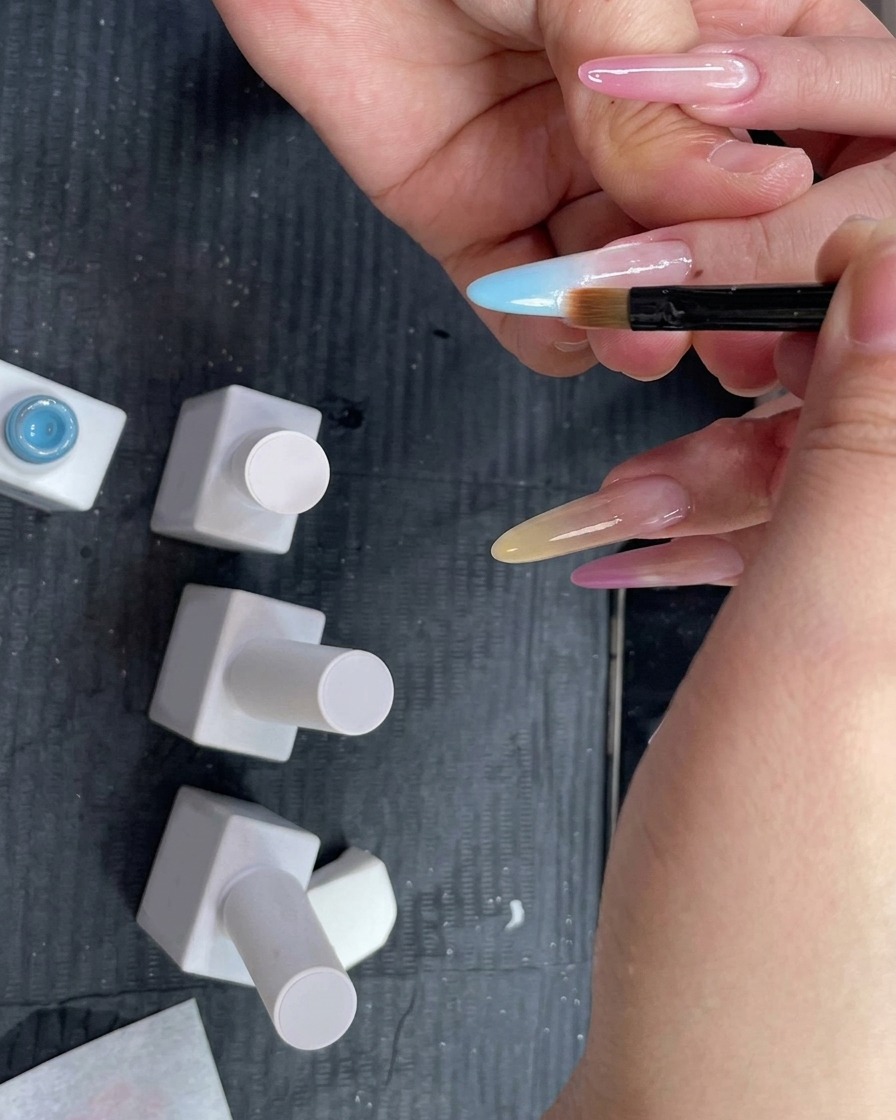 Un pinceau à dégradé tenu à plat contre un ongle pour estomper un gel bleu clair vers le bas.