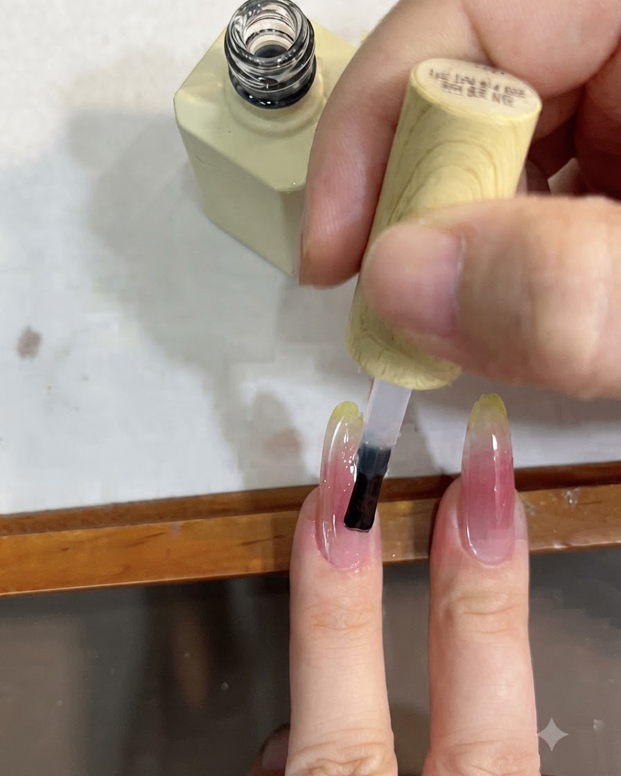 Gros plan dun pinceau appliquant un top coat transparent sur un long ongle au dégradé rose et jaune.