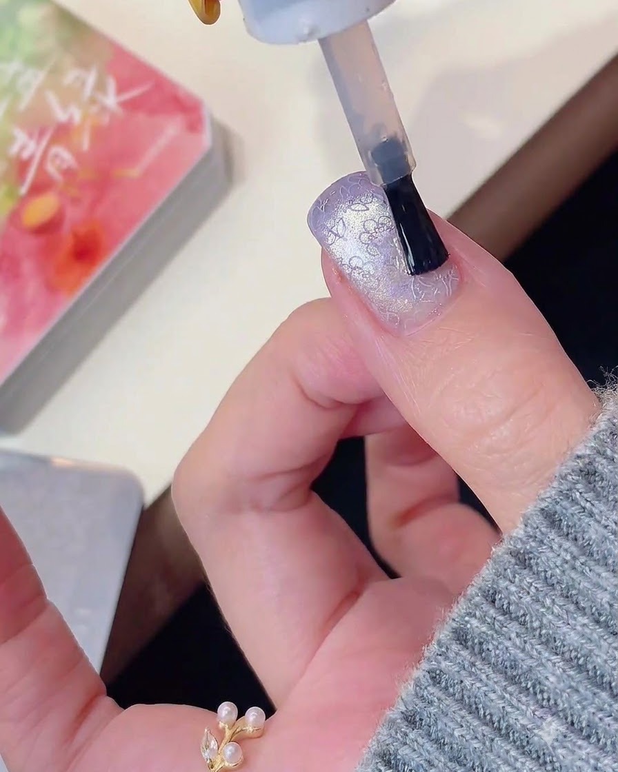 Un pinceau à ongles appliquant un top coat sur un ongle effet œil de chat violet orné dun motif.