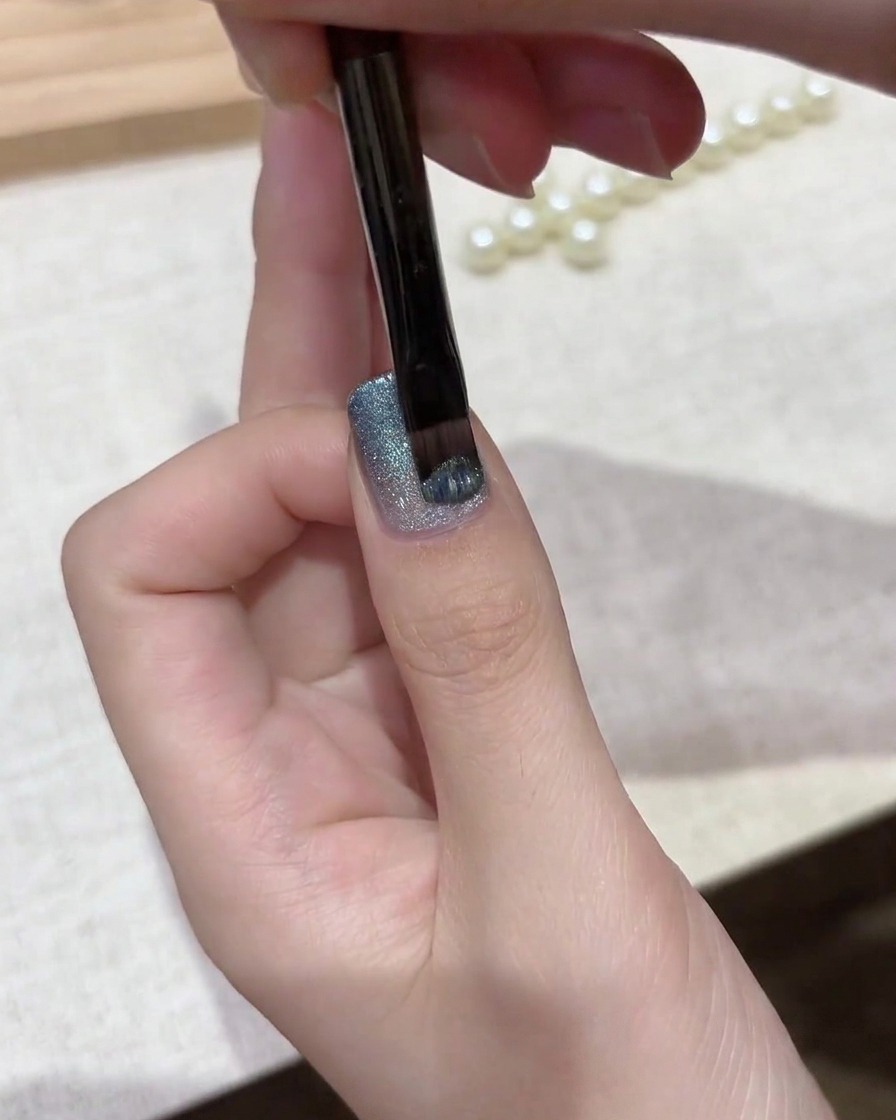 Un pinceau à gel appliquant une couche de vernis cat eye magnétique argent-bleu chatoyant sur une base en dégradé.