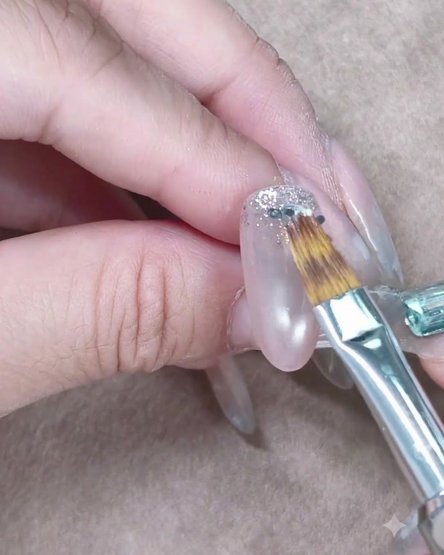 Un petit pinceau à ongle étirant délicatement les paillettes argentées vers le bas sur un ongle en gel transparent.