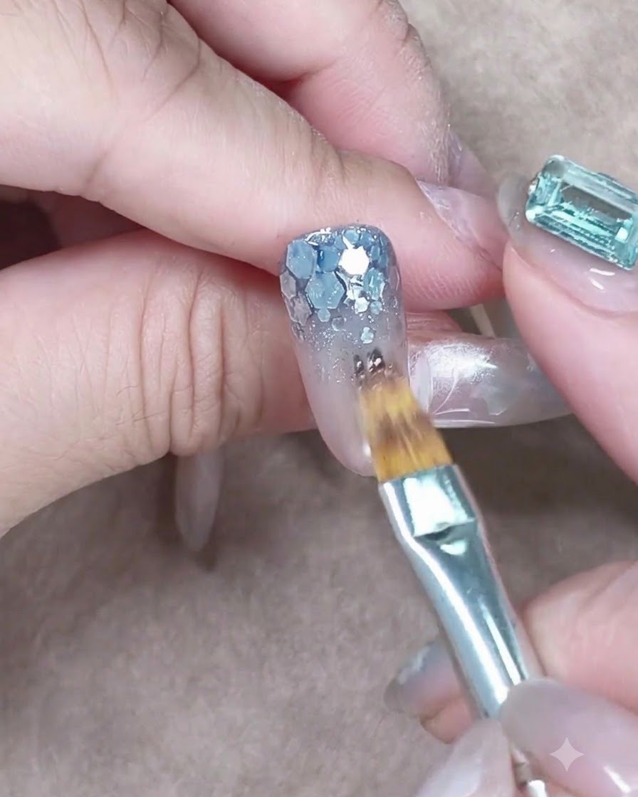 Un pinceau plat appliquant par touches un gel bleu diaphane sur les sequins argentés épais à la base de longle.