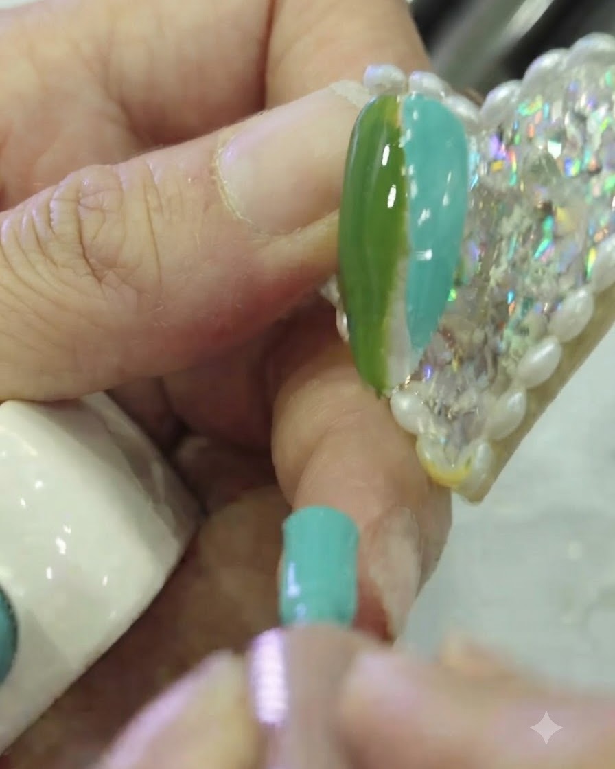 Gros plan dun pinceau plat pour nail art tapotant la frontière entre le vernis gel vert foncé et le vernis gel teal sur un ongle.