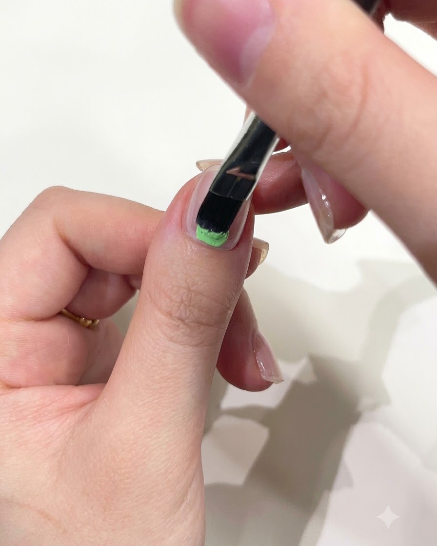 Gros plan dune main utilisant un pinceau gel plat pour appliquer du gel solide vert menthe sur la base dun ongle de pouce.