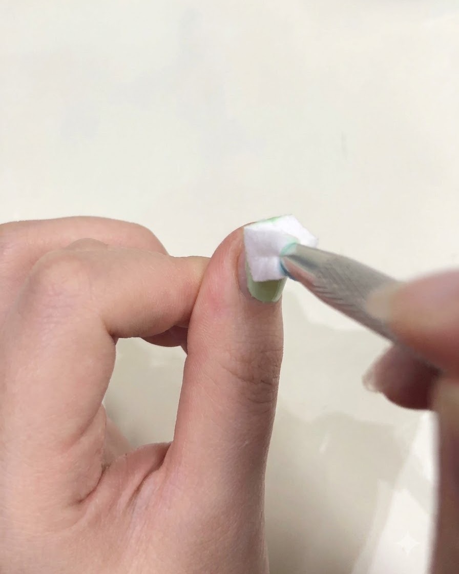 Gros plan dune main tenant une pince à épiler avec une petite éponge blanche tamponnant du gel vert sur un ongle.