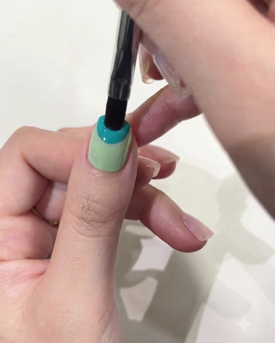 Gros plan dun pinceau gel plat appliquant du gel solide bleu pastel sur la moitié supérieure dun ongle de pouce.