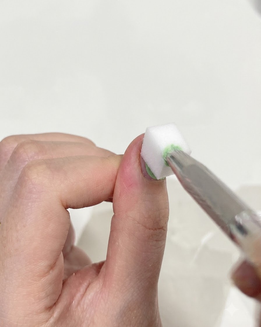 Gros plan dune pince à épiler tenant une petite éponge de maquillage blanche, tapotant délicatement la surface dun ongle pour mélanger les gels vert et bleu.