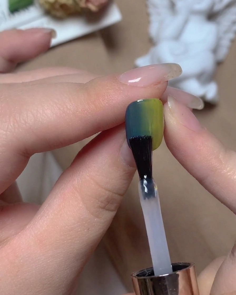 Un pinceau appliquant un top coat transparent sur un ongle dégradé bleu et vert sous un éclairage de studio.