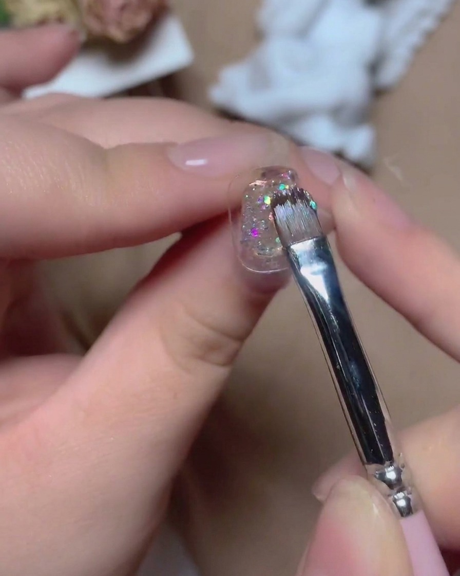 Un pinceau fin appliquant un gel pailleté multicolore étincelant sur un ongle aux reflets vibrants.