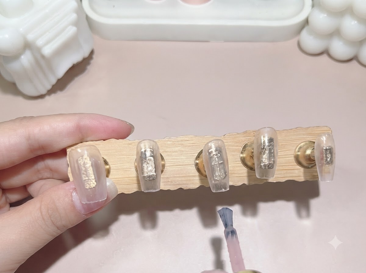 Un pinceau à vernis gel appliquant une seconde couche de rose translucide laiteux sur des capsules dongles fixées sur un présentoir en bois.