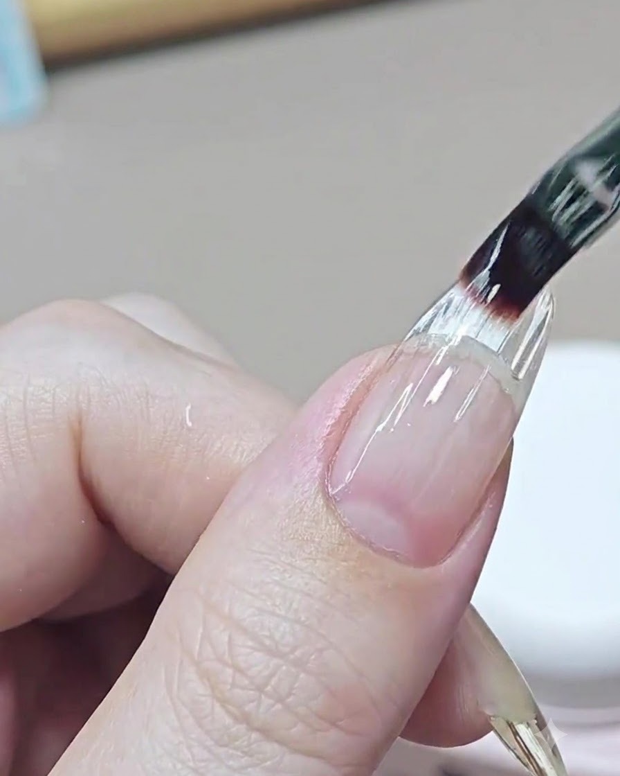 Un pinceau fin applique une couche de gel de construction transparent autonivelant sur la zone de transition dun ongle rallongé.