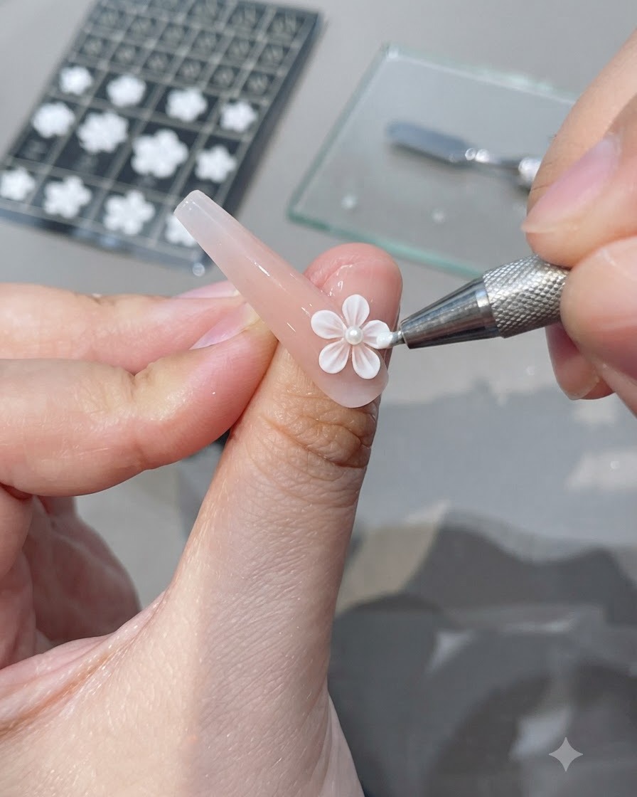 Un outil de précision plaquant un embellissement de fleur 3D blanche sur un ongle en gel rose.