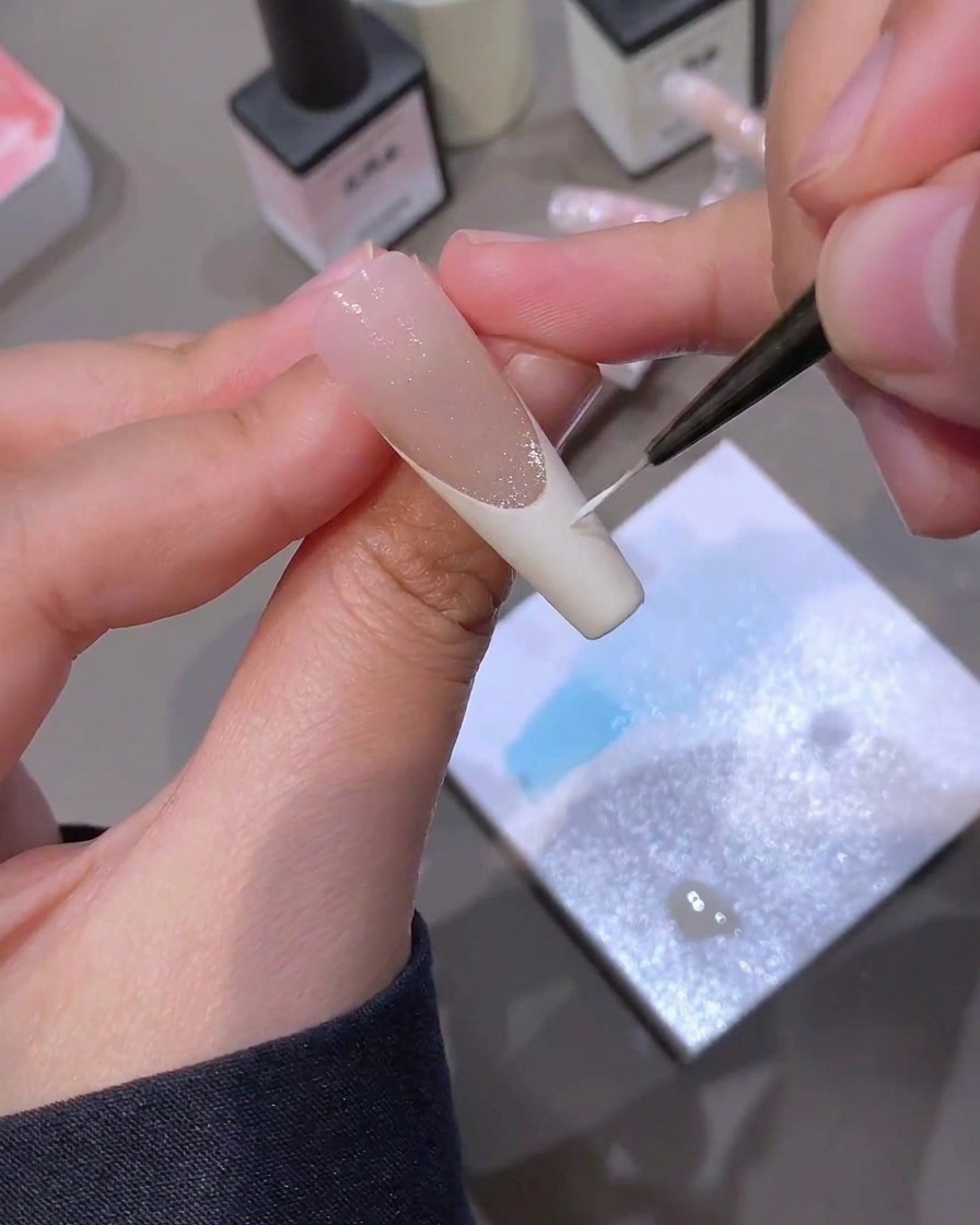 Un pinceau fin de nail art peignant méticuleusement une ligne de pointe French blanche sur un ongle étendu.