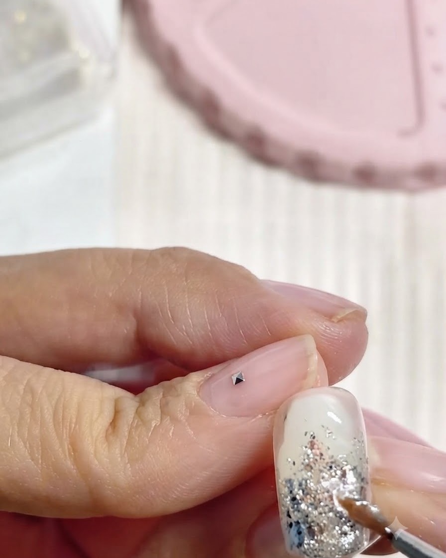 Un stylo en silicone plaquant un petit charm cristal carré sur un doigt à côté dun ongle décoré.