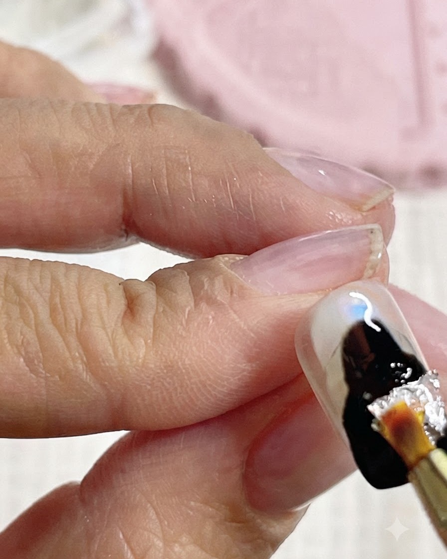 Un pinceau déposant des fragments de feuille dargent sur la zone en gel noir dun ongle.