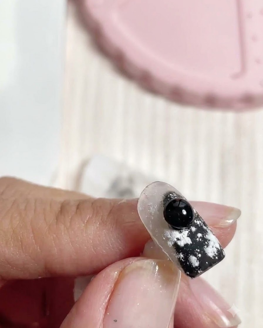 Un pinceau appliquant du vernis noir sur un élément sphérique 3D fixé sur un ongle décoré.