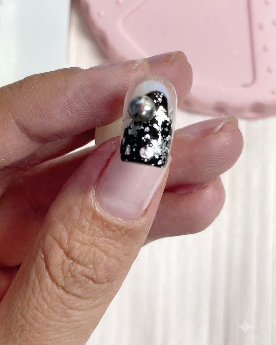 Un ongle court terminé arborant un fond noir brillant avec des éclats de feuille dargent et une boule métallique argentée 3D polie.