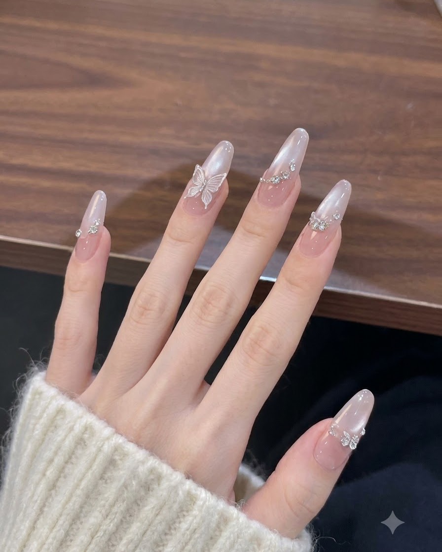 Main aux ongles longs en forme damande fléchissant doucement contre une surface en bois chaleureuse, mettant en valeur un fini rose nud nacré et des charms papillons délicats.