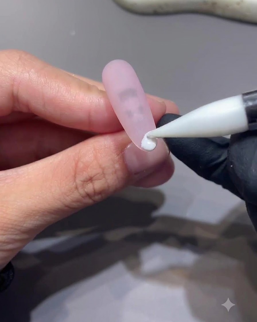 Un outil à pointe fine plaquant une petite goutte de gel blanc sur le bout dun ongle rose mat.