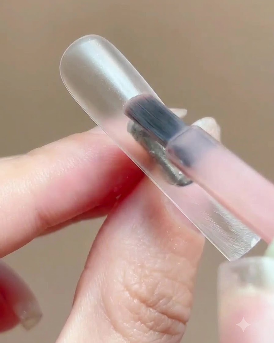 Un pinceau appliquant une fine couche de vernis gel rose translucide sur un présentoir à ongles transparent.