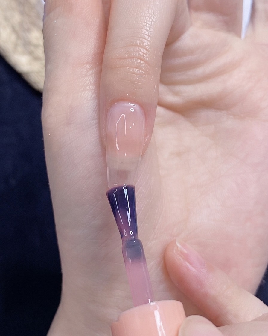 Un pinceau appliquant du vernis gel rose translucide sur un ongle nu.