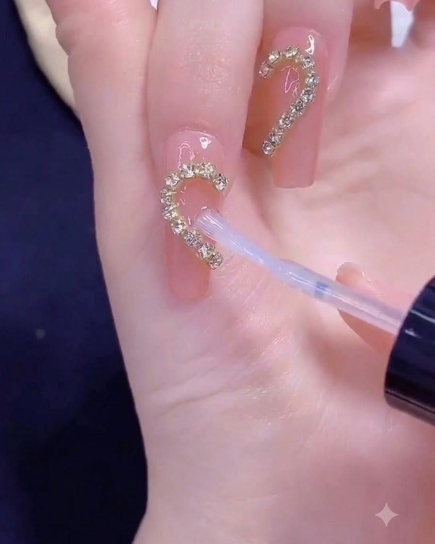 Un pinceau appliquant un top coat transparent sur une bande de chaîne dorée disposée en forme de demi-cœur sur un ongle.