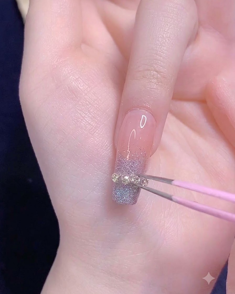Une pince à épiler rose posant une petite bande de chaîne métallique à lhorizontale sur le bout en dégradé dun ongle de lannulaire.