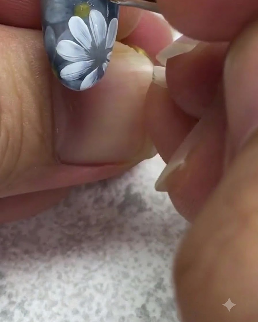 Un pinceau de détail fin appliquant un point de gel jaune vif au centre dune marguerite blanche peinte sur un ongle gris.