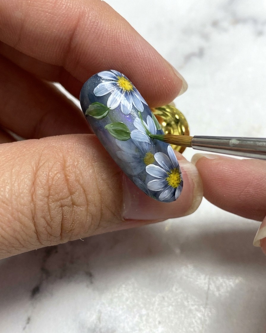 Un pinceau de nail art ajoutant une petite feuille verte à côté des détails floraux blancs sur une base dongle gris.