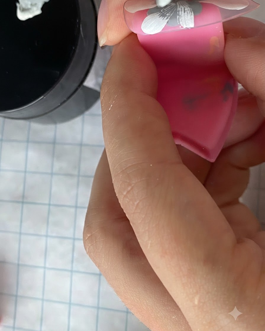 Un pinceau fin remplissant un groupe de pétales de marguerite blanche peints sur un ongle monté sur support rose.
