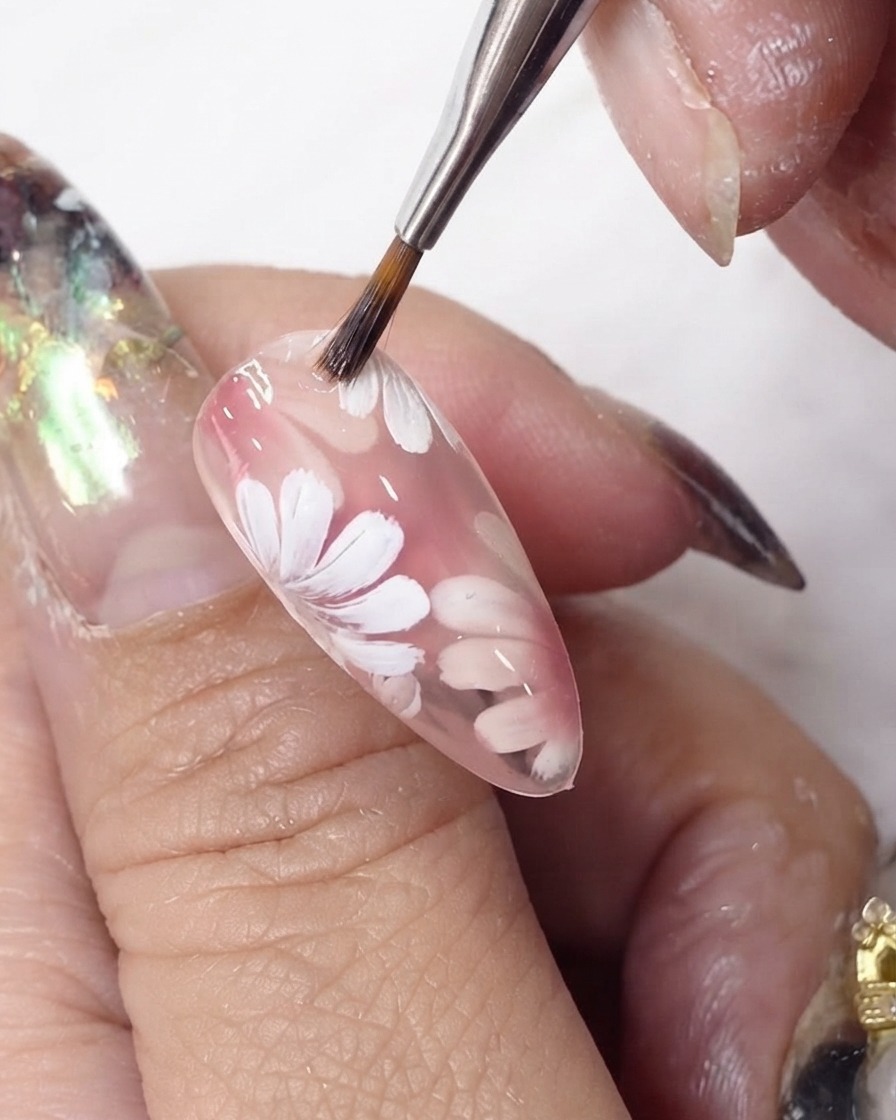 Un pinceau à gel faisant glisser une couche de vernis rose diaphane sur des fleurs blanches peintes sur un ongle en amande.