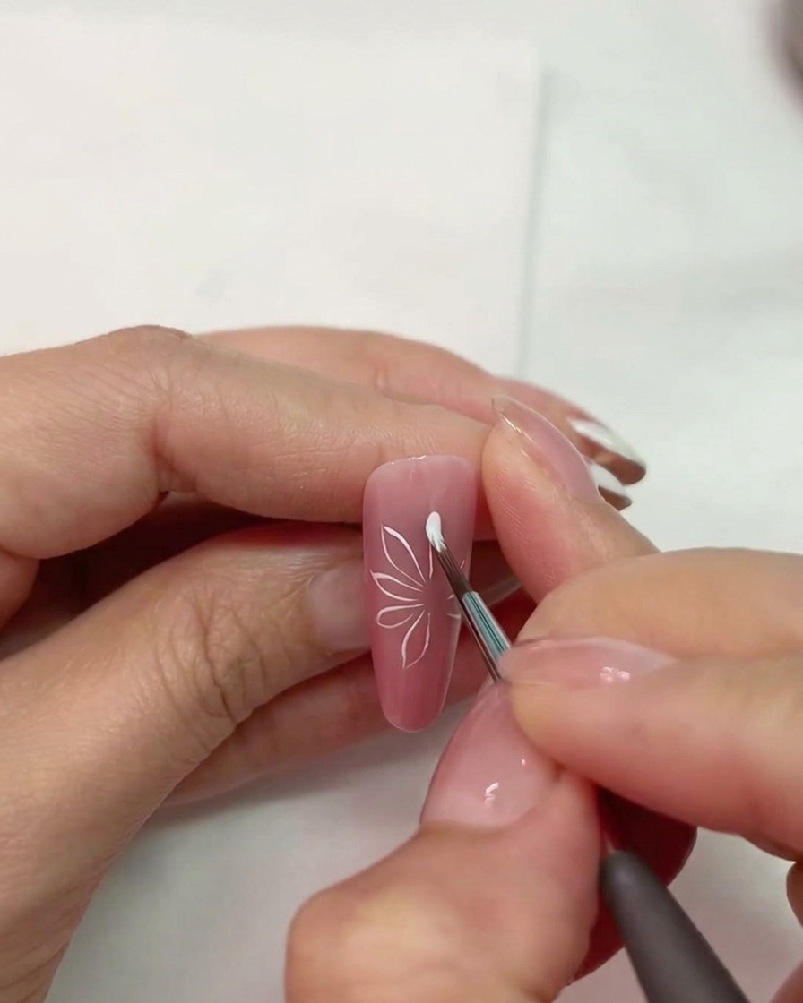 Gros plan dun pinceau à ongles dessinant des formes de pétales blancs sur un ongle rose mat.