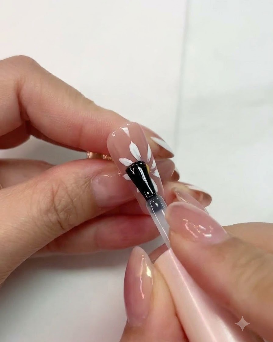 Un pinceau appliquant un top coat mat sur un design de marguerite blanche catalysé sur un ongle rose.