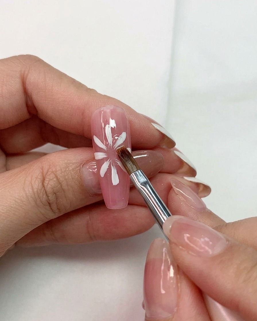 Une main tenant un pinceau polyvalent façonnant des pétales blancs sur une base rose sur un ongle.