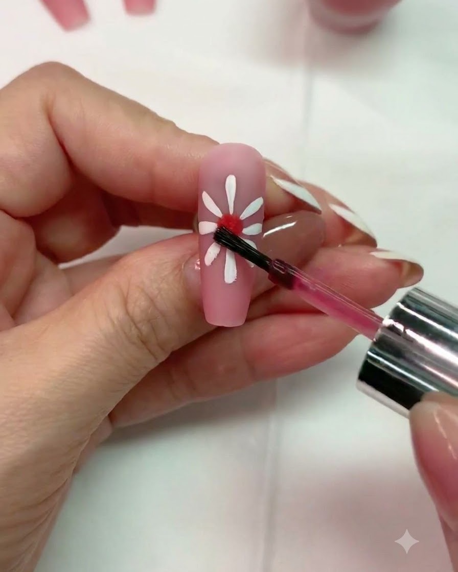 Un pinceau appliquant une petite goutte de gel blooming rouge au centre dun design de marguerite blanche sur un ongle rose mat.