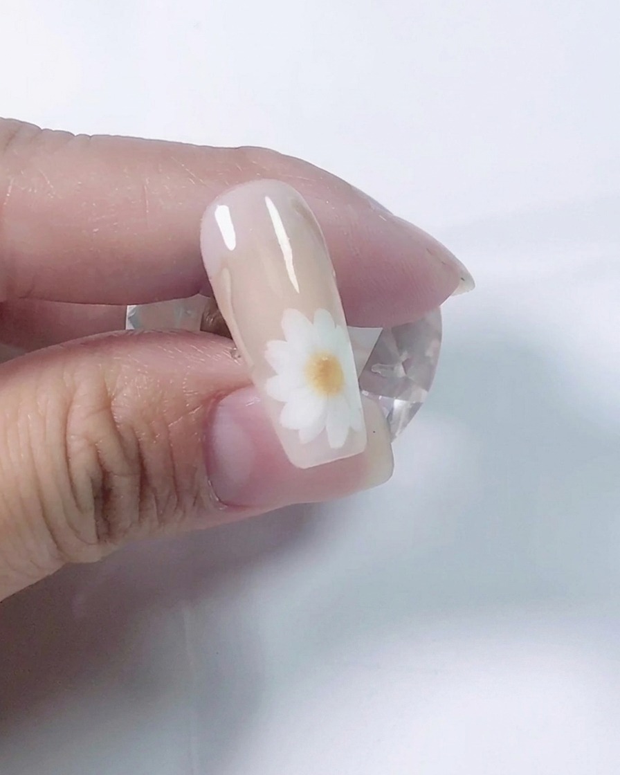 Gros plan dune prothésiste ongulaire utilisant un pinceau fin pour peindre des pétales de marguerite blancs sur un ongle dentraînement nude.