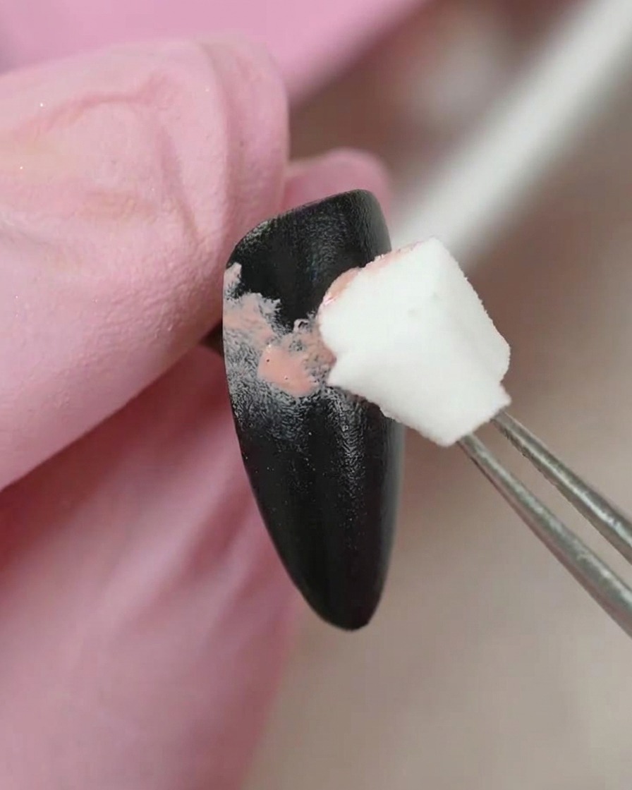 Une paire de brucelles tenant un petit applicateur éponge, tamponnant délicatement du gel de couleur rose sur la surface dun ongle peint en noir.