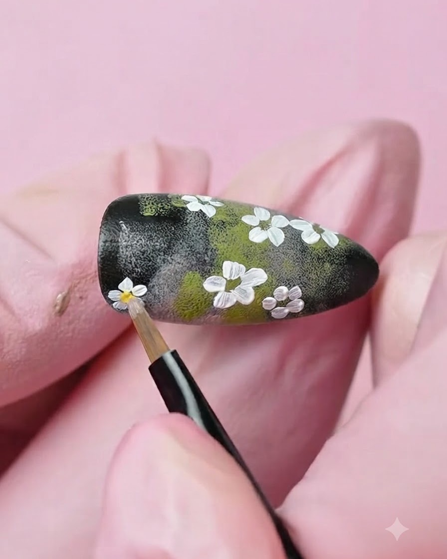 Un pinceau de détail fin appliquant une goutte de gel jaune au centre dune fleur blanche peinte sur longle.