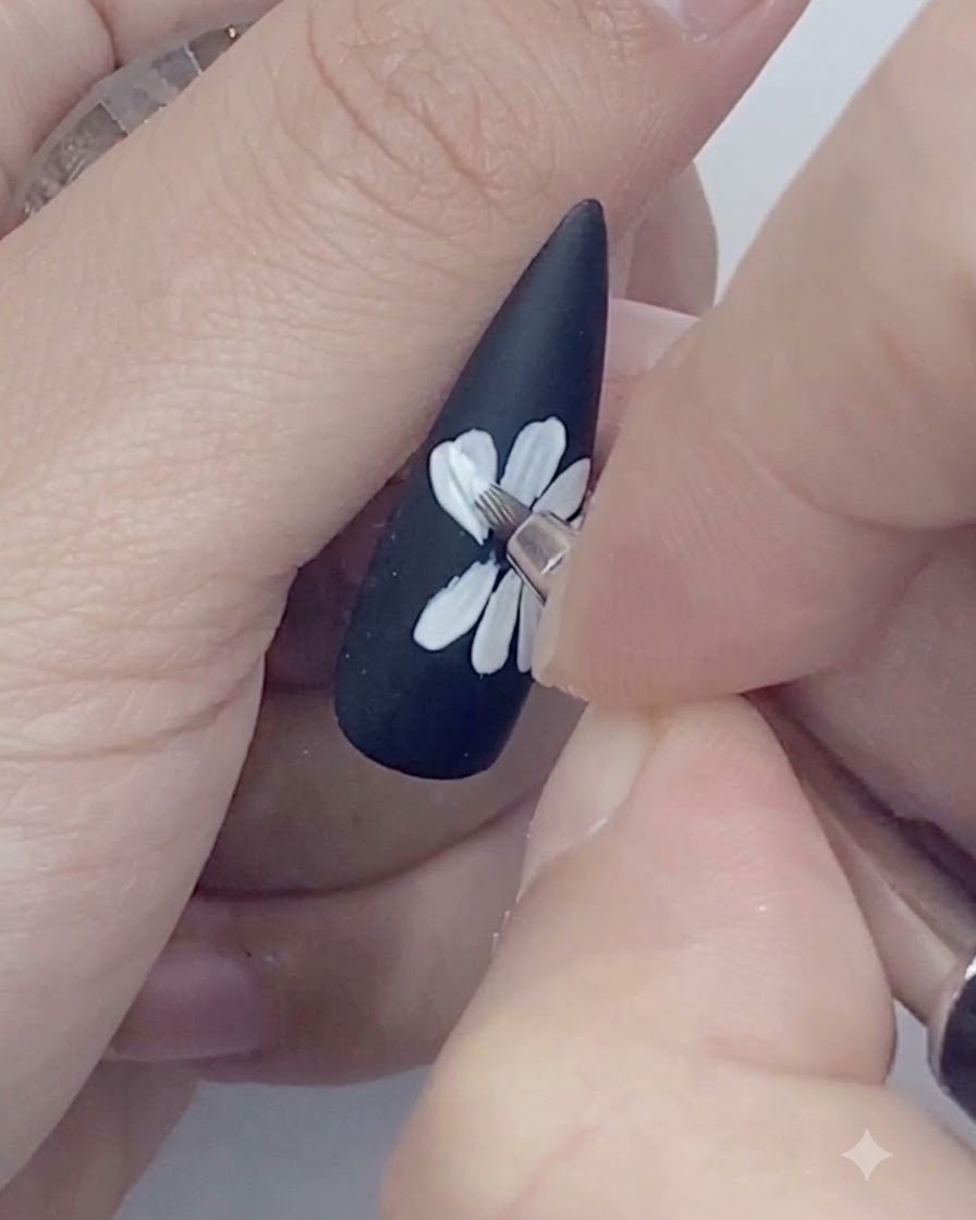 Un pinceau fin traceur dessinant le contour extérieur des pétales de marguerite blanche sur un ongle noir.