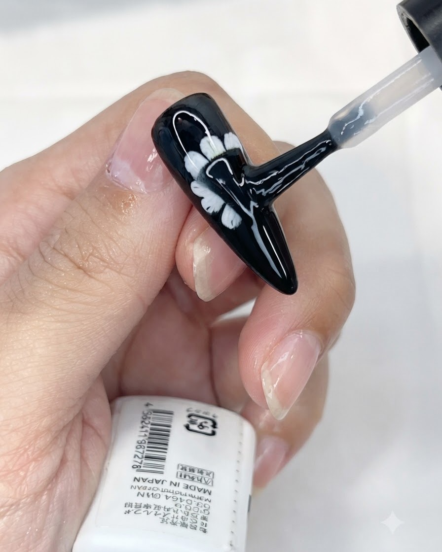 Un pinceau appliquant une couche de top coat mat sur un ongle noir décoré dune marguerite blanche.